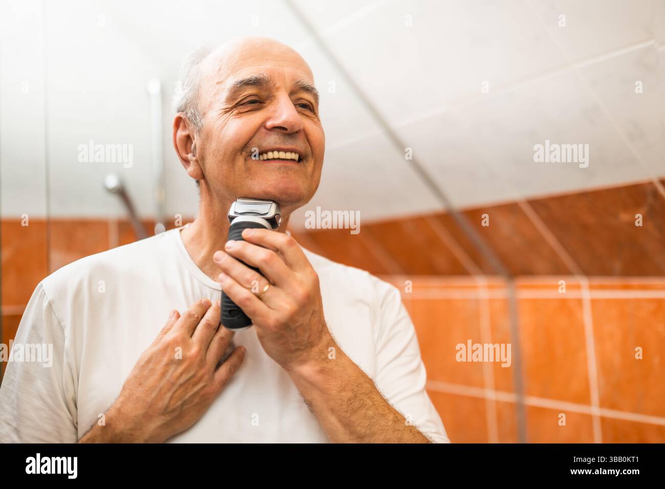 Portrait d'homme senior heureux regardant son reflet tout en rasant la barbe avec rasoir électrique dans la salle de bain. Banque D'Images