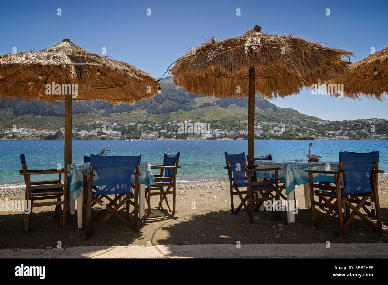 Deux tables, chaises et parasols de paille sur la plage au bord de la mer Égée sur l'île de Telendos en Grèce par une journée ensoleillée. Kalymnos est la rive opposée. Banque D'Images
