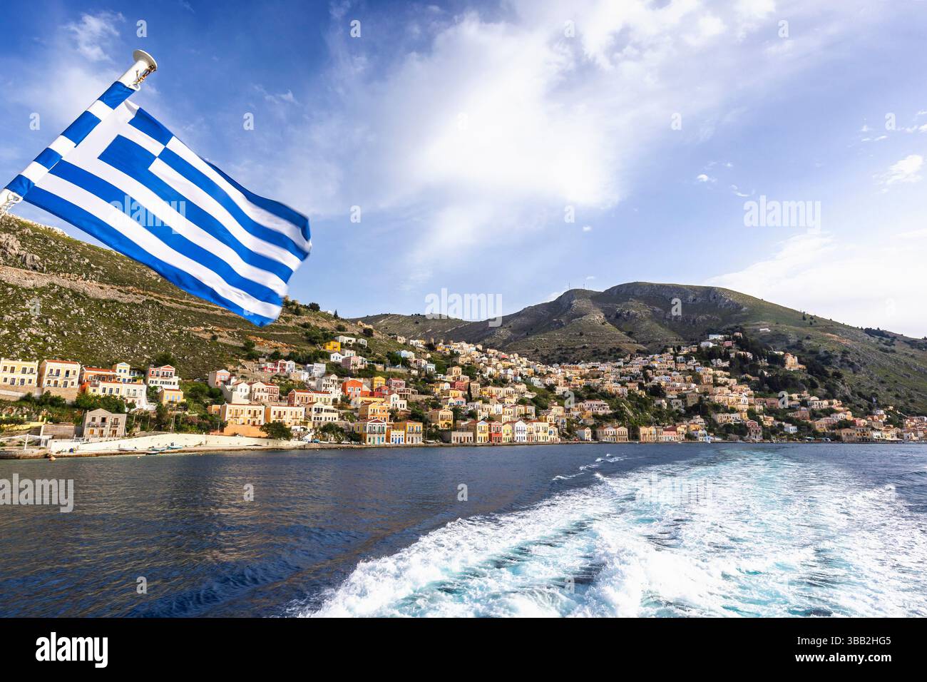 Bâtiments colorés sur une colline dans la ville de Symi par la mer Égée sur Symi (Simi, Syme) île en Grèce par une journée ensoleillée. Le drapeau grec est au premier plan. Banque D'Images