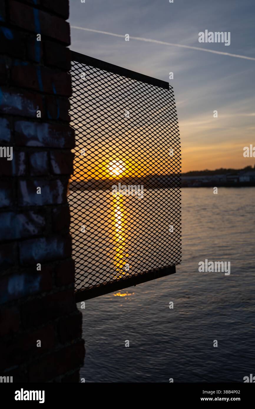 Coucher de soleil se reflétant sur la surface de l'eau, vu à travers un treillis métallique attaché à un mur de briques pendant l'heure dorée Banque D'Images