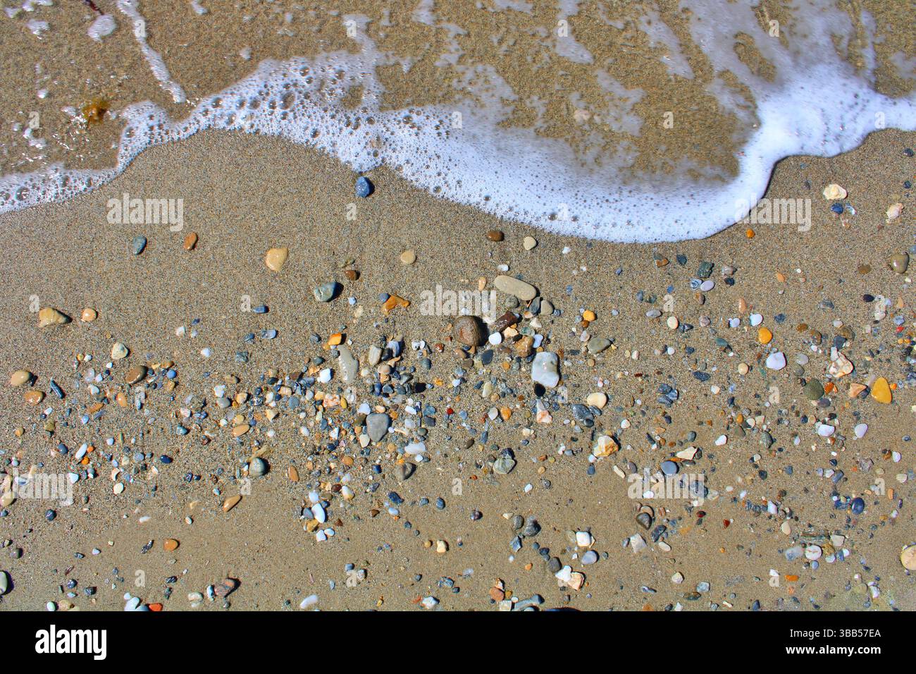 Petites pierres colorées sur une plage de sable avec de l'écume marine des vagues, capturées d'en haut par une journée d'été ensoleillée. Banque D'Images