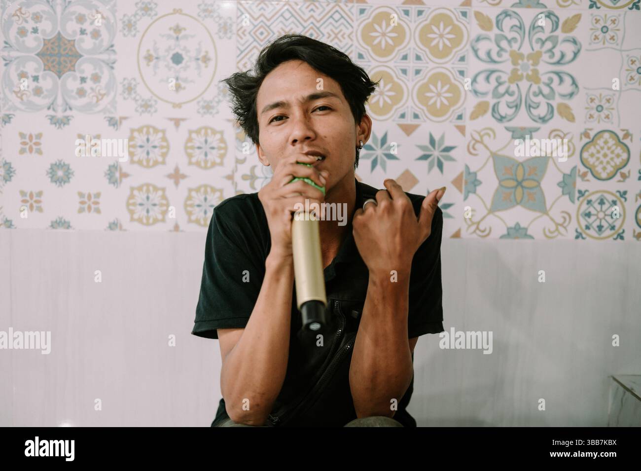 Jeune homme asiatique chantant avec microphone contre des carreaux à motifs. Des performances captivantes avec une touche de style personnel. Banque D'Images