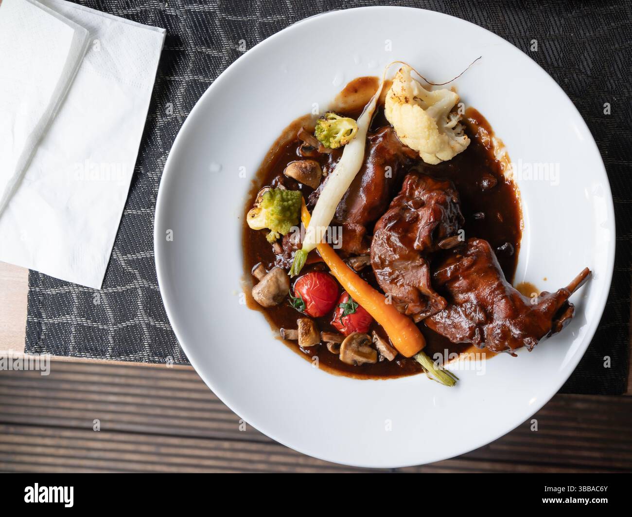 Sur une table en bois, se trouve une assiette blanche classique magnifiquement surmontée d'une portion copieuse de viande et d'une variété de légumes frais, créant un col Banque D'Images