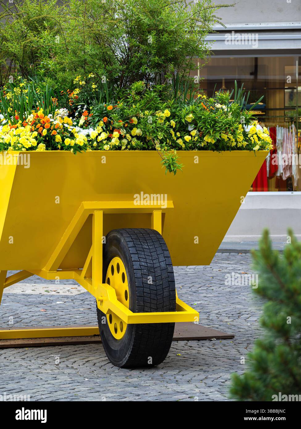 Brouette jaune éclatante remplie de fleurs colorées et de verdure, située contre une rue pavée. La scène est animée et accueillante, mettant en valeur Banque D'Images