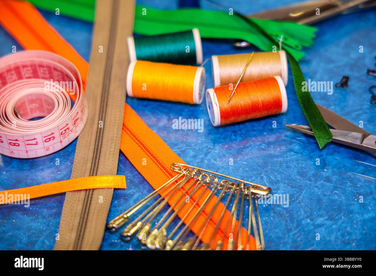 Matériaux de couture brillants sur la surface de travail bleue, y compris des bobines de fil, des fermetures éclair orange et verte, des épingles de sécurité, des ciseaux et un ruban à mesurer rose Banque D'Images