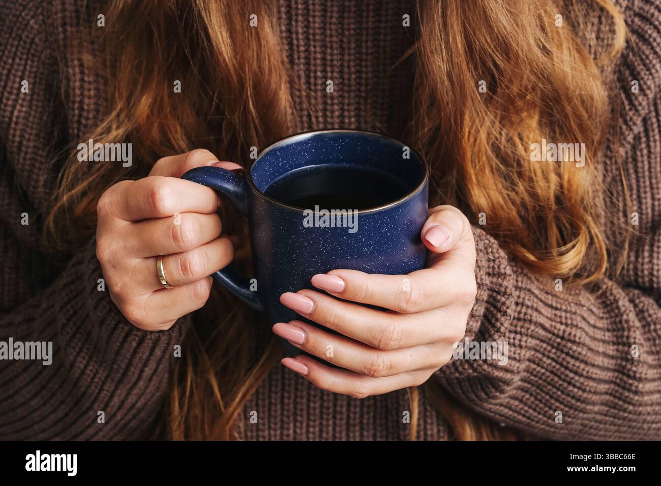 Femme tenant une tasse à café. Boisson chaude dans une tasse bleue. Pull confortable et cheveux longs. Boisson relaxante du matin. Mains féminines avec manucure. Banque D'Images