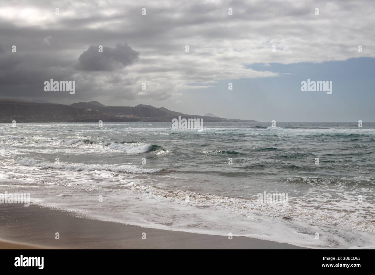 Plage de sable avec vagues de l'océan Atlantique. Montagnes à l'horizon. Ciel bleu avec des nuages blancs. Las Palmas de Gran Canaria, Îles Canaries, Espagne. Banque D'Images