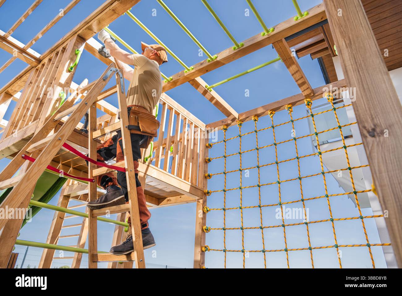 Un travailleur de la construction installe soigneusement des dispositifs de sécurité sur une structure de jeu en bois dans une cour arrière résidentielle. La lumière du soleil met en valeur le travail Banque D'Images