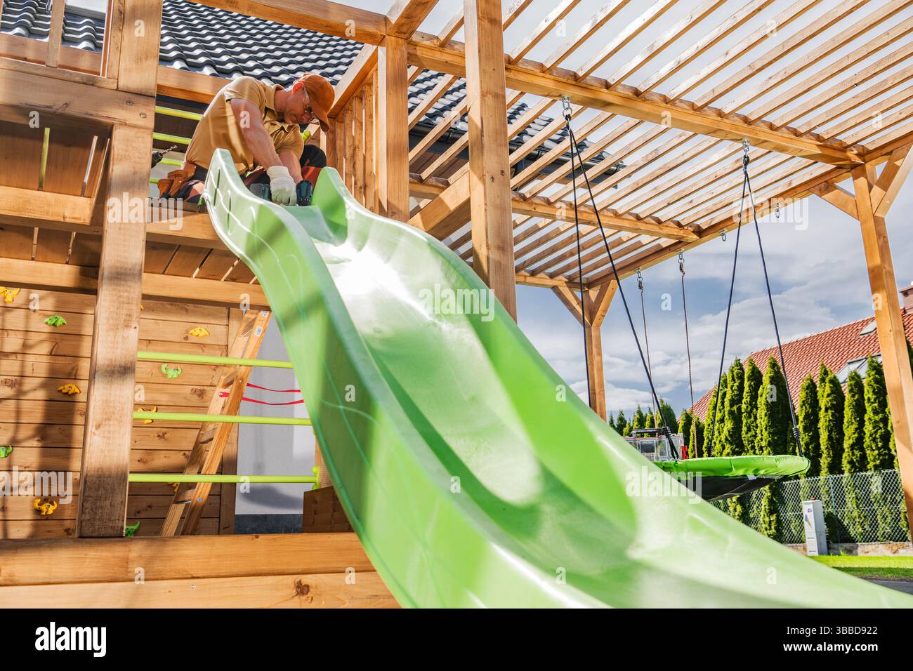 Un homme installe soigneusement une toboggan vert vif sur une structure de jeu en bois dans une cour arrière résidentielle. L'aire de jeux dispose de poutres en bois, escalade Banque D'Images