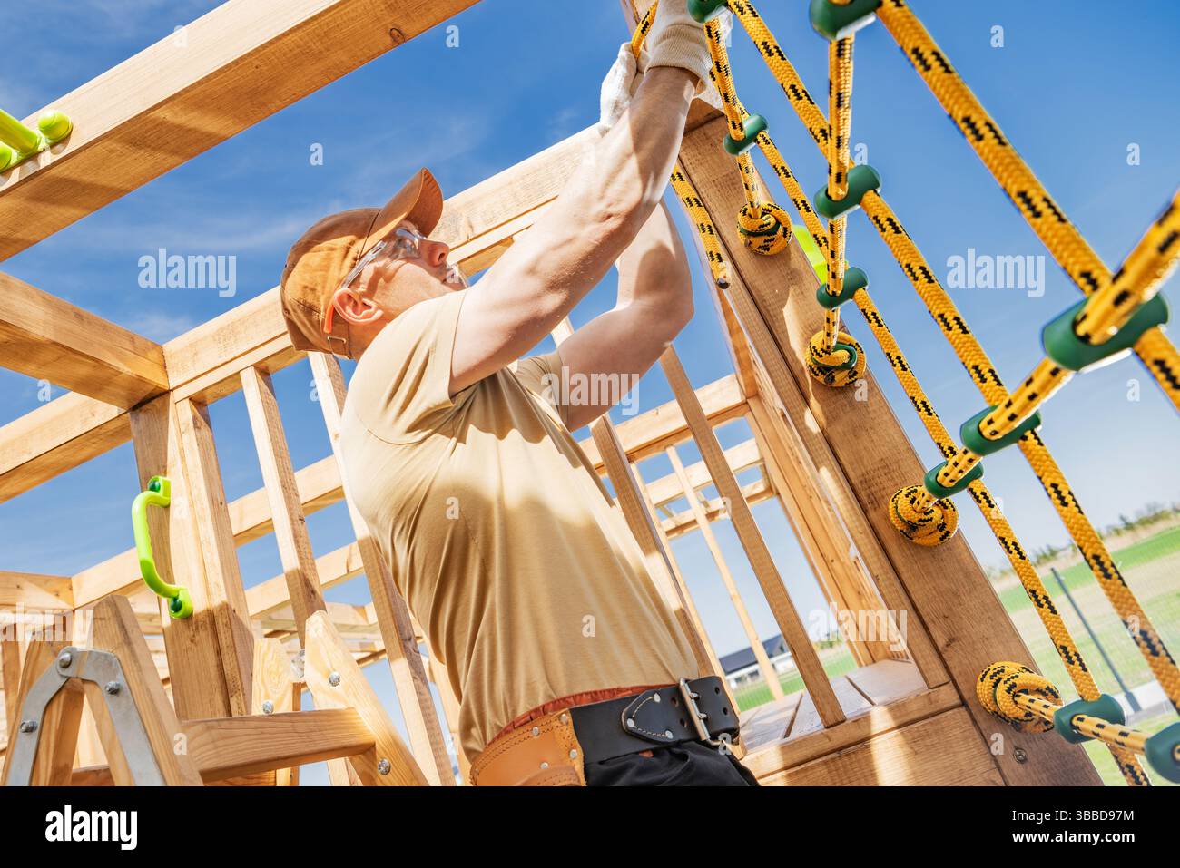 Dans un parc animé sous un ciel bleu clair, un travailleur qualifié assemble des cordes d'escalade pour une structure de jeu en bois. Les matériaux de construction, inclu Banque D'Images