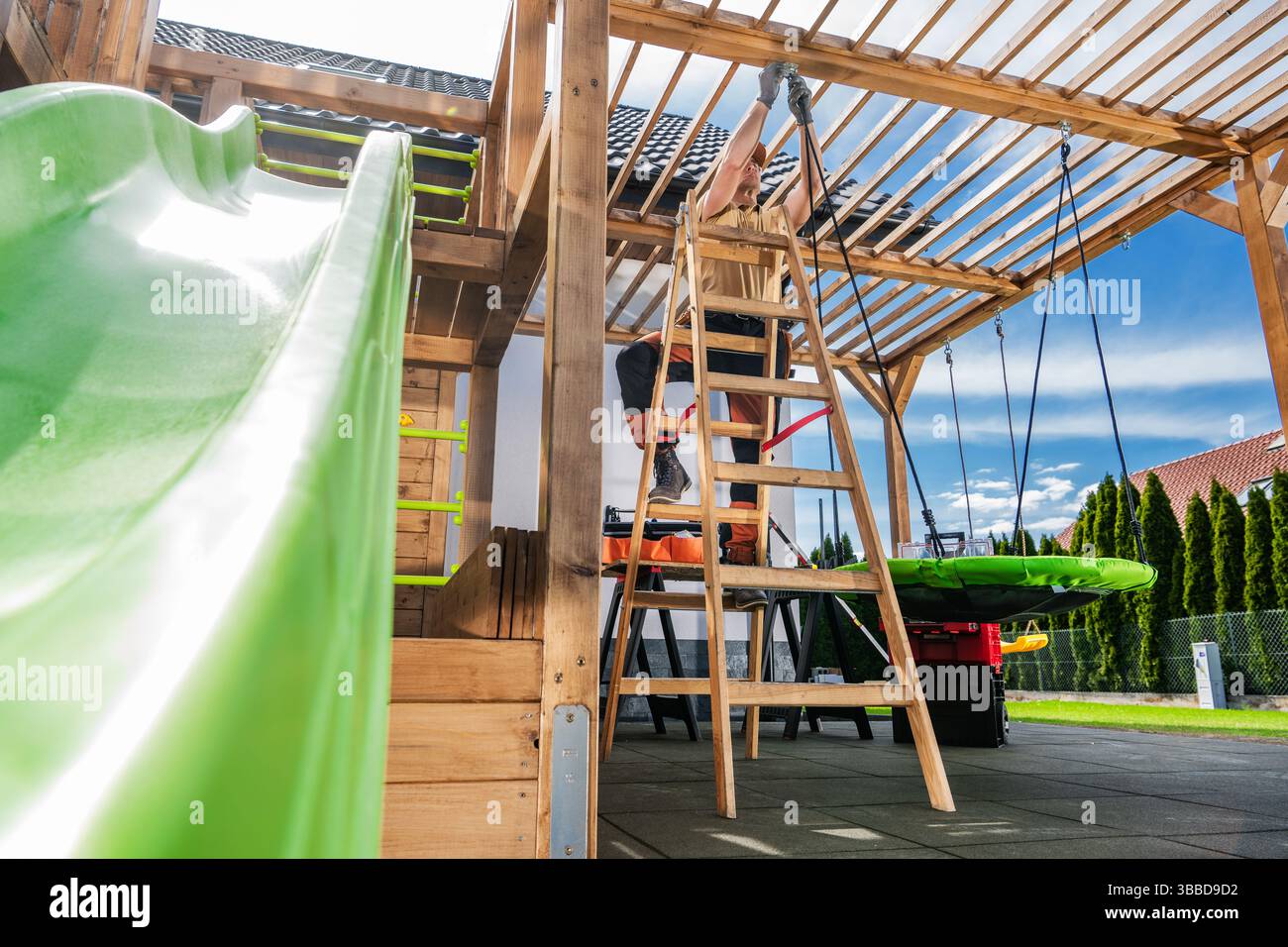 Un enfant aime glisser sur un toboggan vert vif tandis qu'un parent travaille à la construction d'une structure de jeu en bois dans une cour arrière spacieuse. Le soleil brille b Banque D'Images