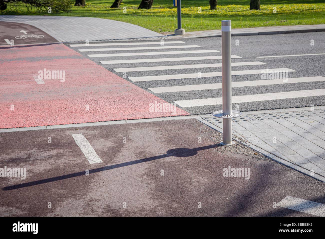 piste cyclable rouge traversant une traversée zèbre dans un cadre urbain moderne. La scène présente un poteau métallique projetant une ombre, un trottoir adjacent et un vert Banque D'Images