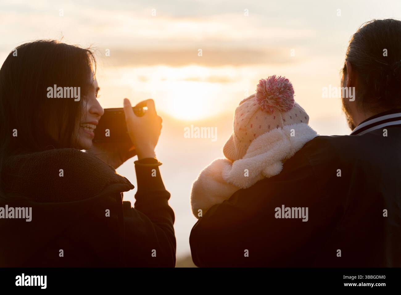 Femme souriante prenant la photo du lever du soleil tandis que l'homme porte bébé dans des vêtements chauds. Famille profitant de la belle lumière du matin ensemble. Concept d'amour, memor Banque D'Images