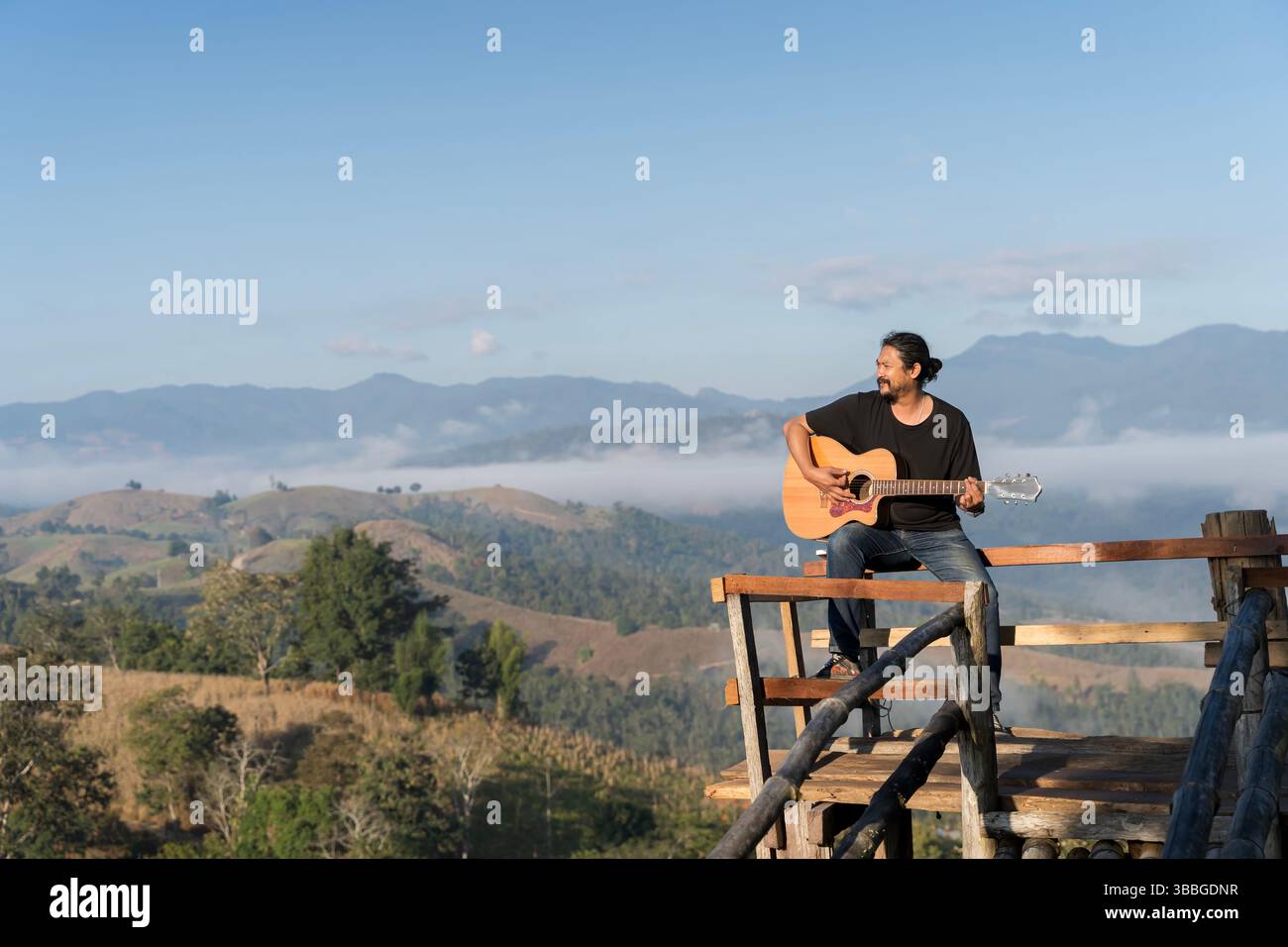 Homme asiatique assis sur une plate-forme en bois jouant de la guitare acoustique, entouré par un paysage de montagne pittoresque et la brume matinale. Moment paisible avec de la musique, na Banque D'Images