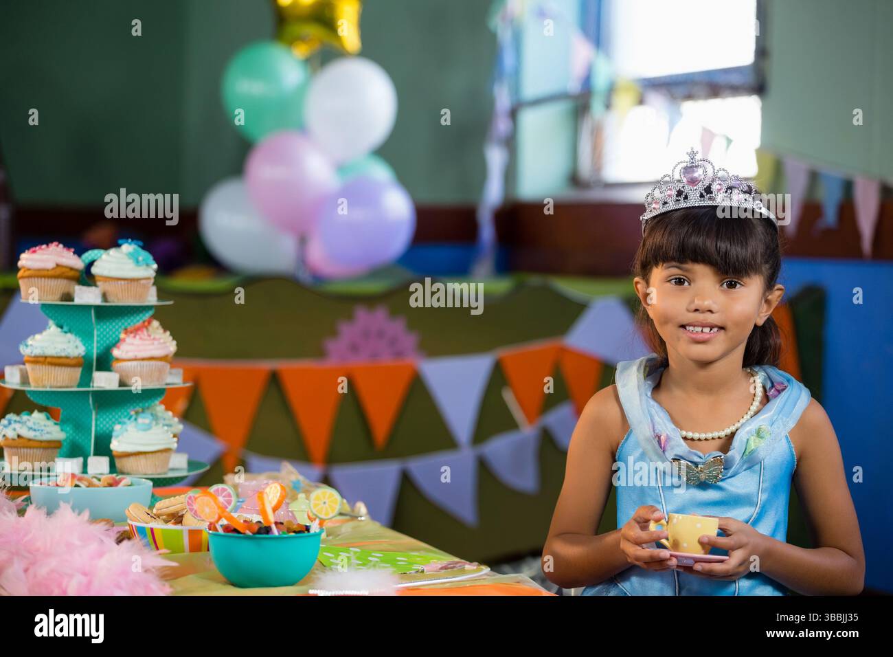 Fille souriante portant la diadème, assise à la table à l'intérieur avec tasse et cupcake stand, espace de copie Banque D'Images