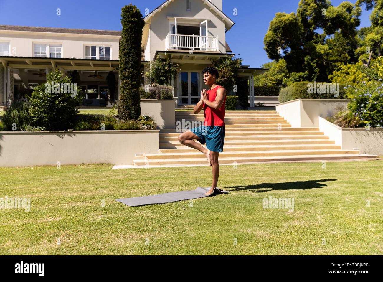 Jeune homme pratiquant la pose d'arbre sur la pelouse devant une maison de deux étages, avec tapis de yoga gris Banque D'Images