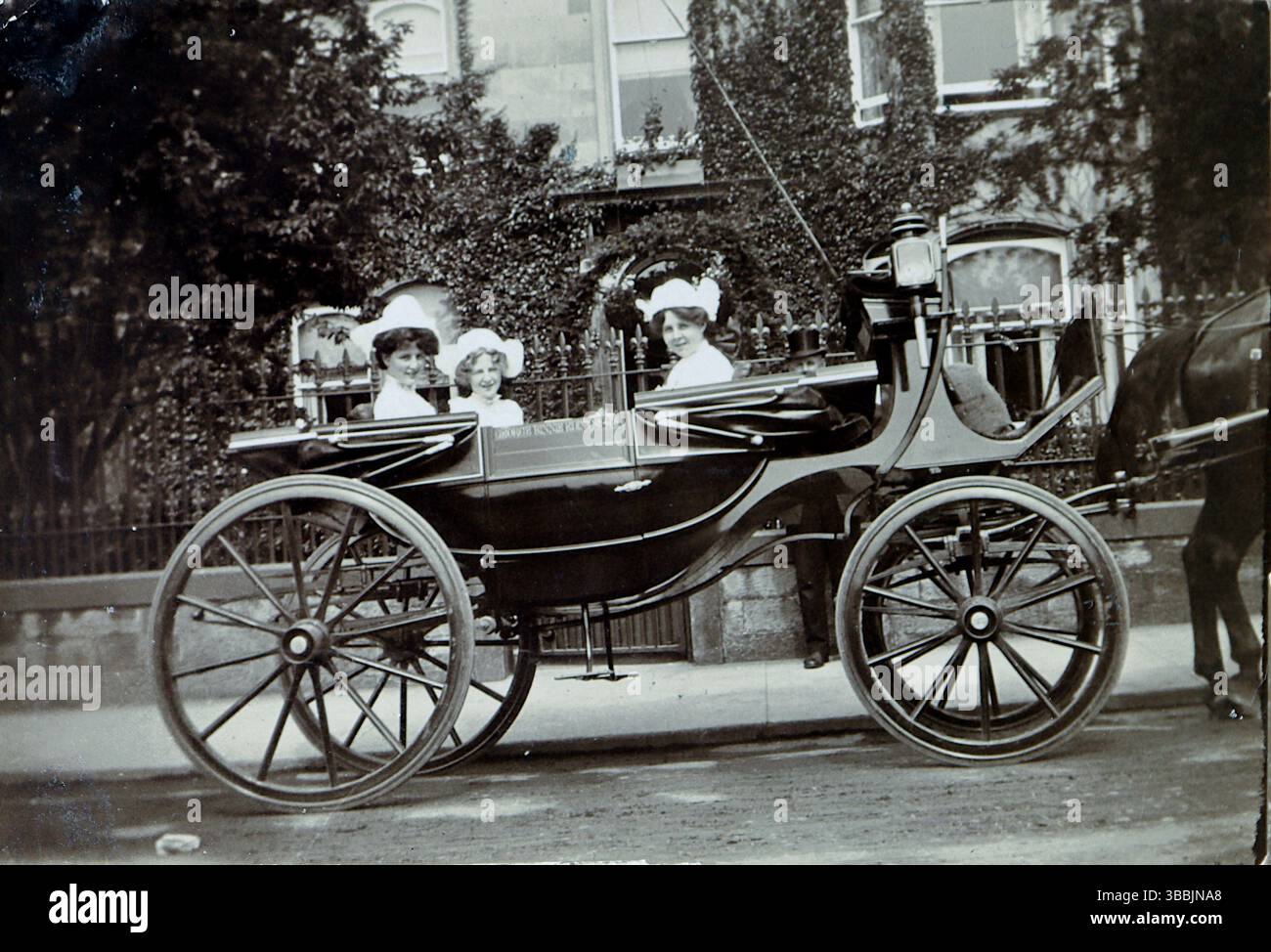 Une calèche tirée par des chevaux avec de grandes roues en bois, un mode de transport qui régissait autrefois les rues. À l'intérieur de la voiture, trois personnes sont assises, toutes portant des chapeaux qui ajoutent au charme démodé de la scène. Le cadre comprend une rue avec une clôture et des bâtiments en arrière-plan. Extrait d'un album victorien et édouardien qui semble avoir été compilé par une personne anonyme. Banque D'Images