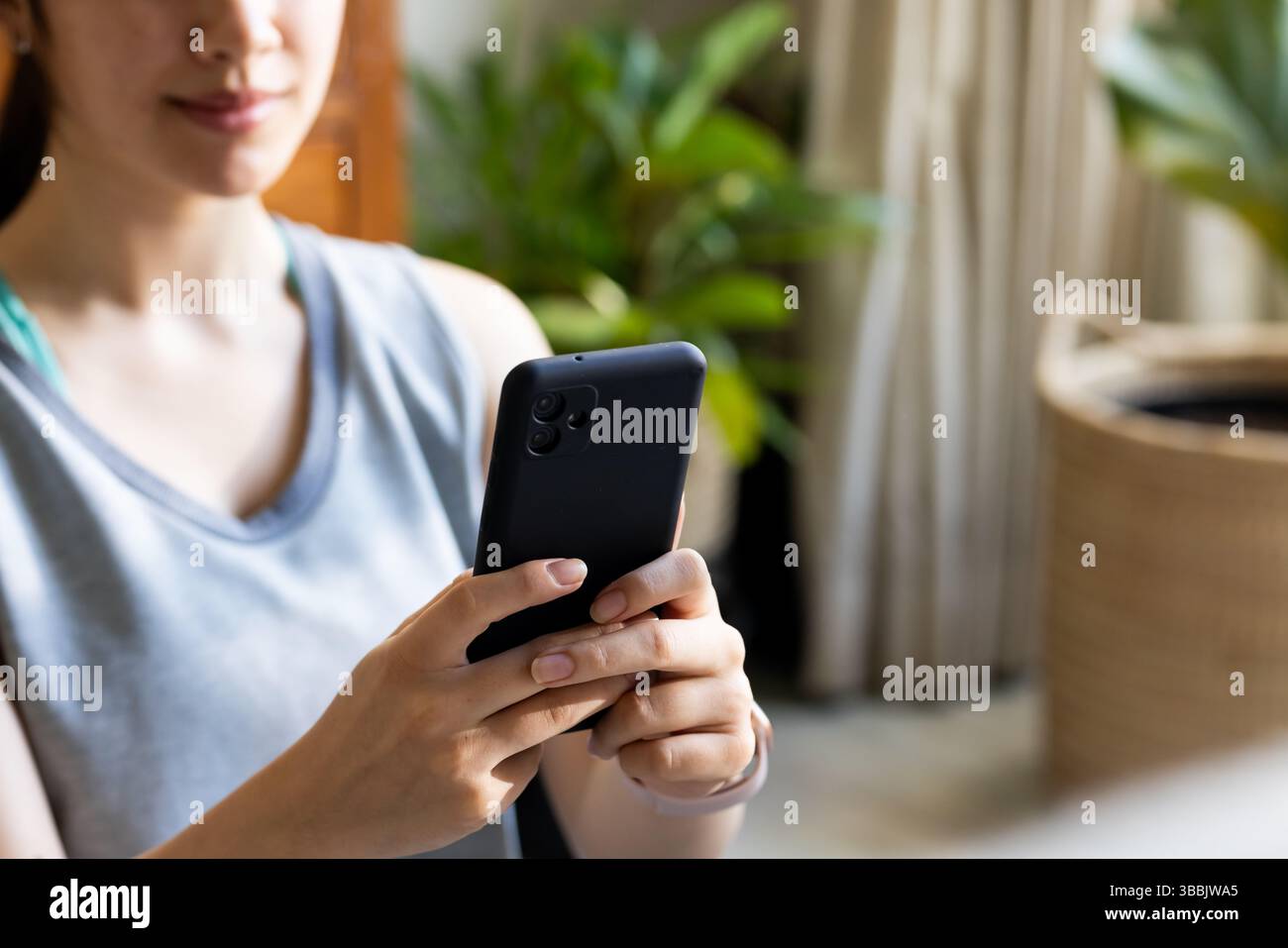 Femme asiatique assise tenant un smartphone noir dans le salon, avec des plantes d'intérieur, espace de copie Banque D'Images