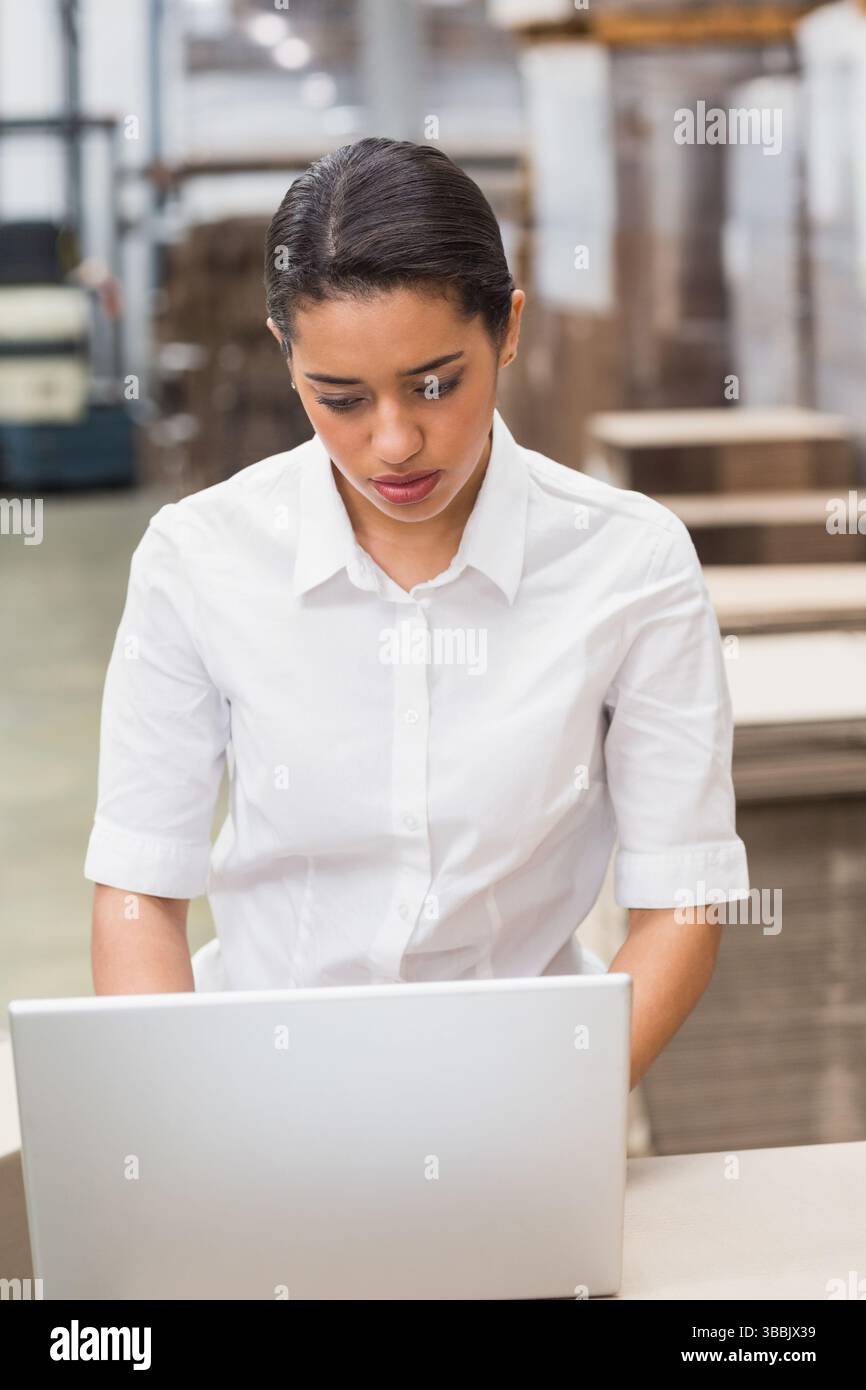 Femme asiatique debout à la table à l'intérieur du grand entrepôt, en utilisant un ordinateur portable argenté près des étagères et des palettes Banque D'Images