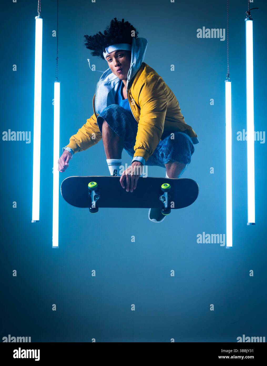 Homme sautant afro-américain en veste jaune tenant skateboard dans un studio bleu, avec des tubes néon Banque D'Images