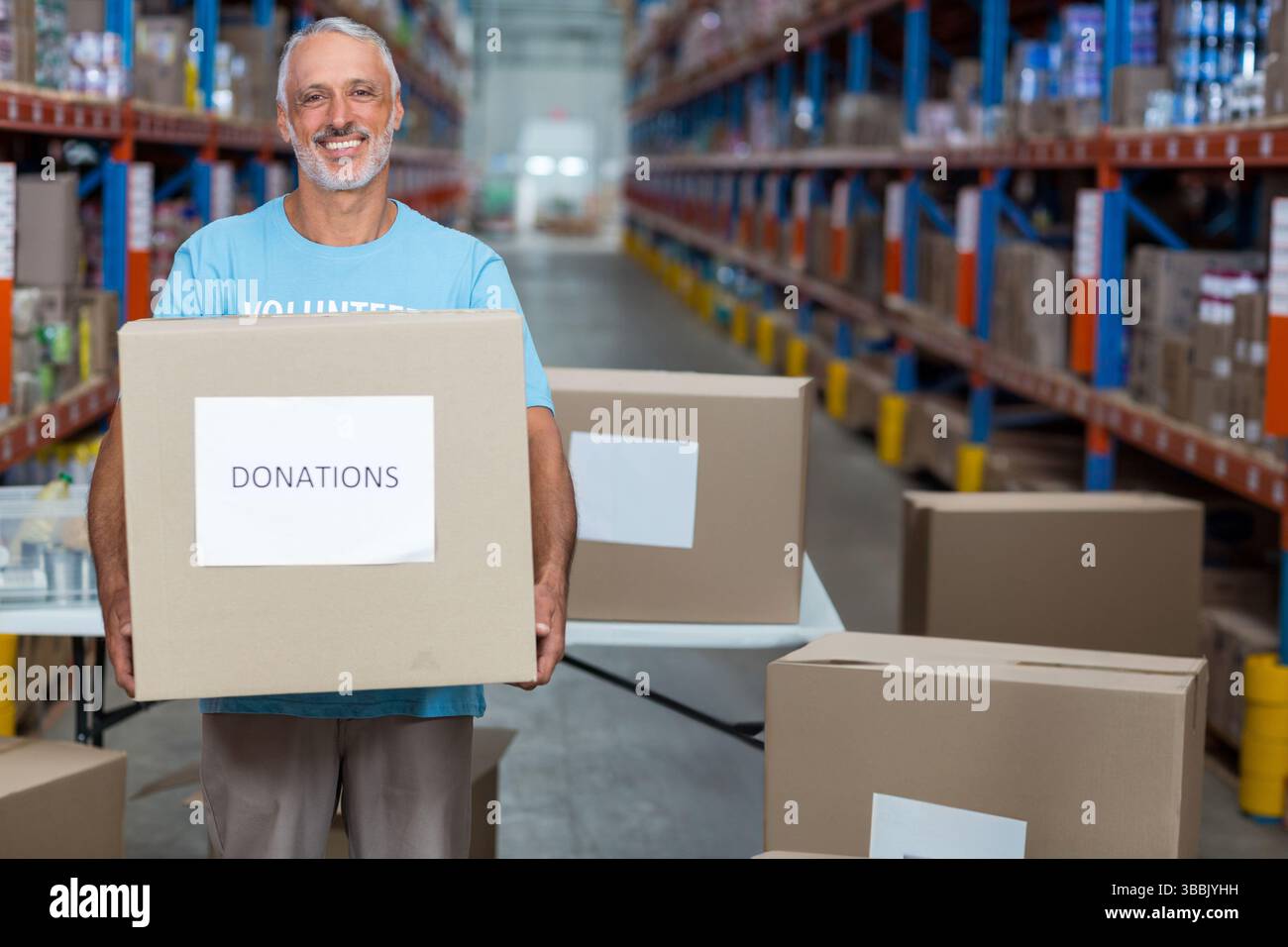 Bénévole de sexe masculin tenant une boîte de dons en carton dans l'allée de l'entrepôt, avec table pliante et étagères Banque D'Images