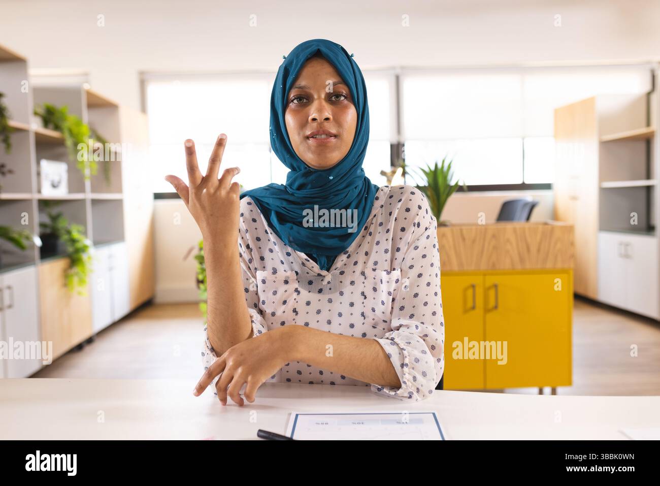 Femme portant le hijab sarcelle montrant trois doigts au bureau avec un stylo de document et une armoire jaune Banque D'Images