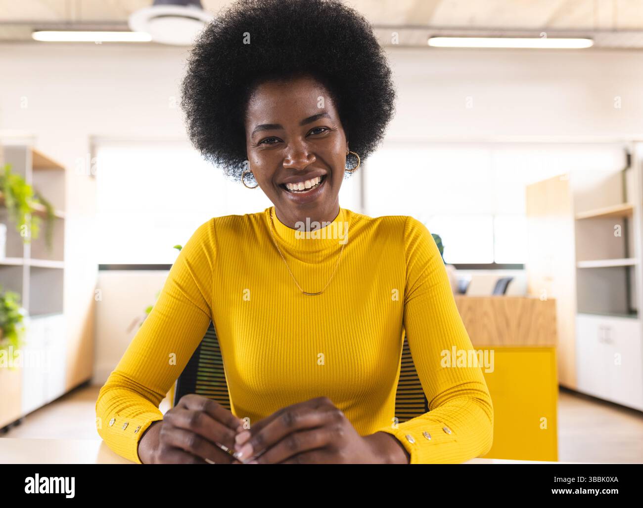 Femme afro-américaine assise au bureau dans un bureau moderne ouvert avec des plantes en pot, souriante Banque D'Images