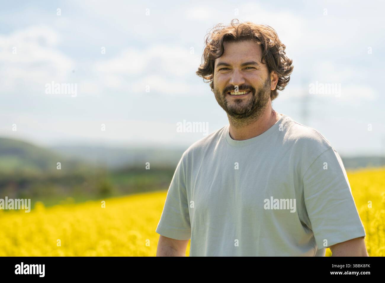 Mignon homme bouclé barbu en T-shirt souriant debout devant un champ de colza fleuri jaune. Concept d'émotions, de loisirs, de nature et de voyage Banque D'Images