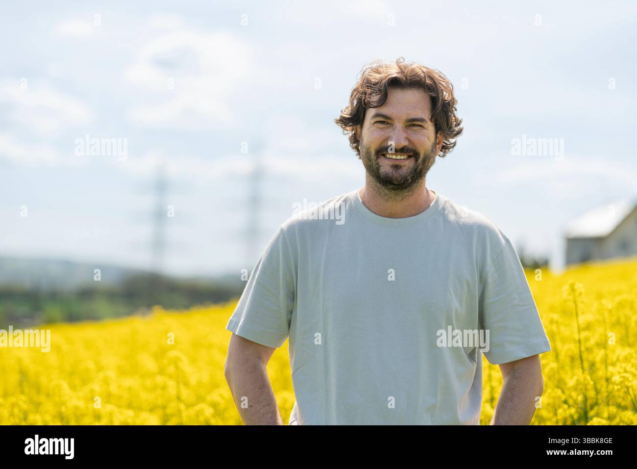 Mignon homme bouclé barbu en T-shirt souriant debout devant un champ de colza fleuri jaune. Concept d'émotions, de loisirs, de nature et de voyage Banque D'Images