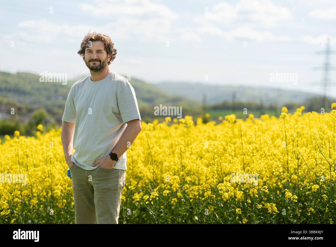 Mignon homme bouclé barbu en T-shirt souriant debout devant un champ de colza fleuri jaune. Concept d'émotions, de loisirs, de nature et de voyage Banque D'Images