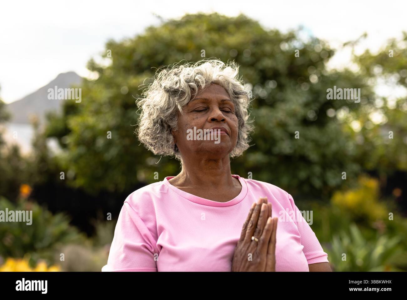 Femme senior méditant dans le parc de jardin avec les mains serrées à la poitrine, exprimant la concentration calme Banque D'Images