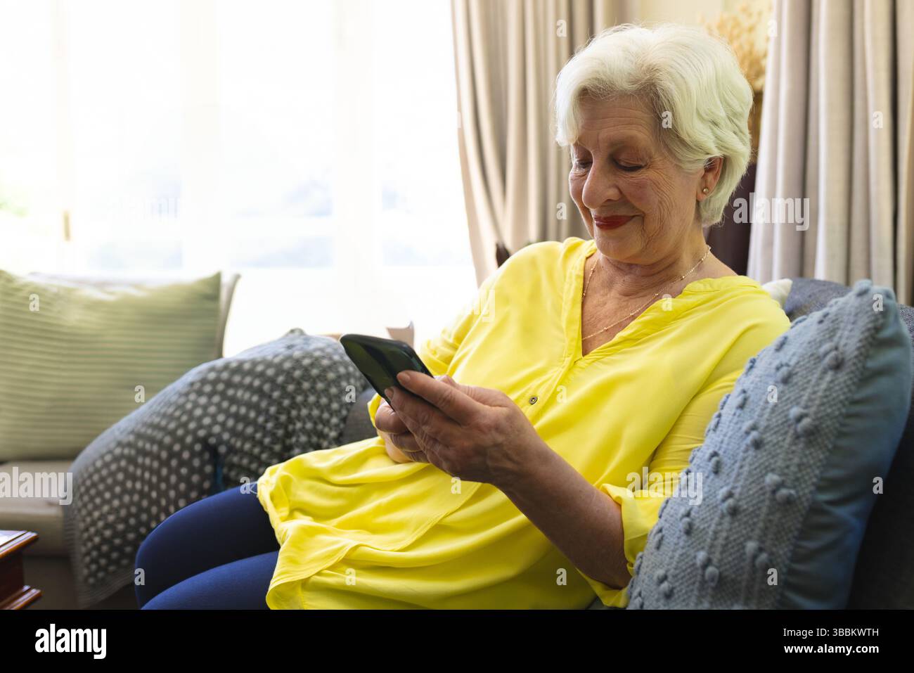 Femme senior assise sur un canapé gris à la maison, à l'aide d'un smartphone avec des oreillers et des rideaux, espace de copie Banque D'Images