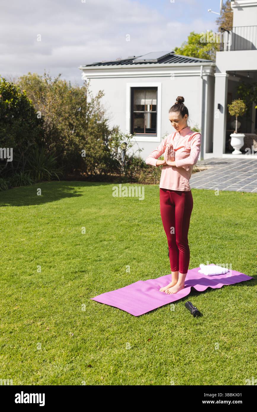 Jeune femme pratiquant le yoga de prière pose sur tapis violet dans la cour arrière de banlieue, avec serviette et bouteille Banque D'Images