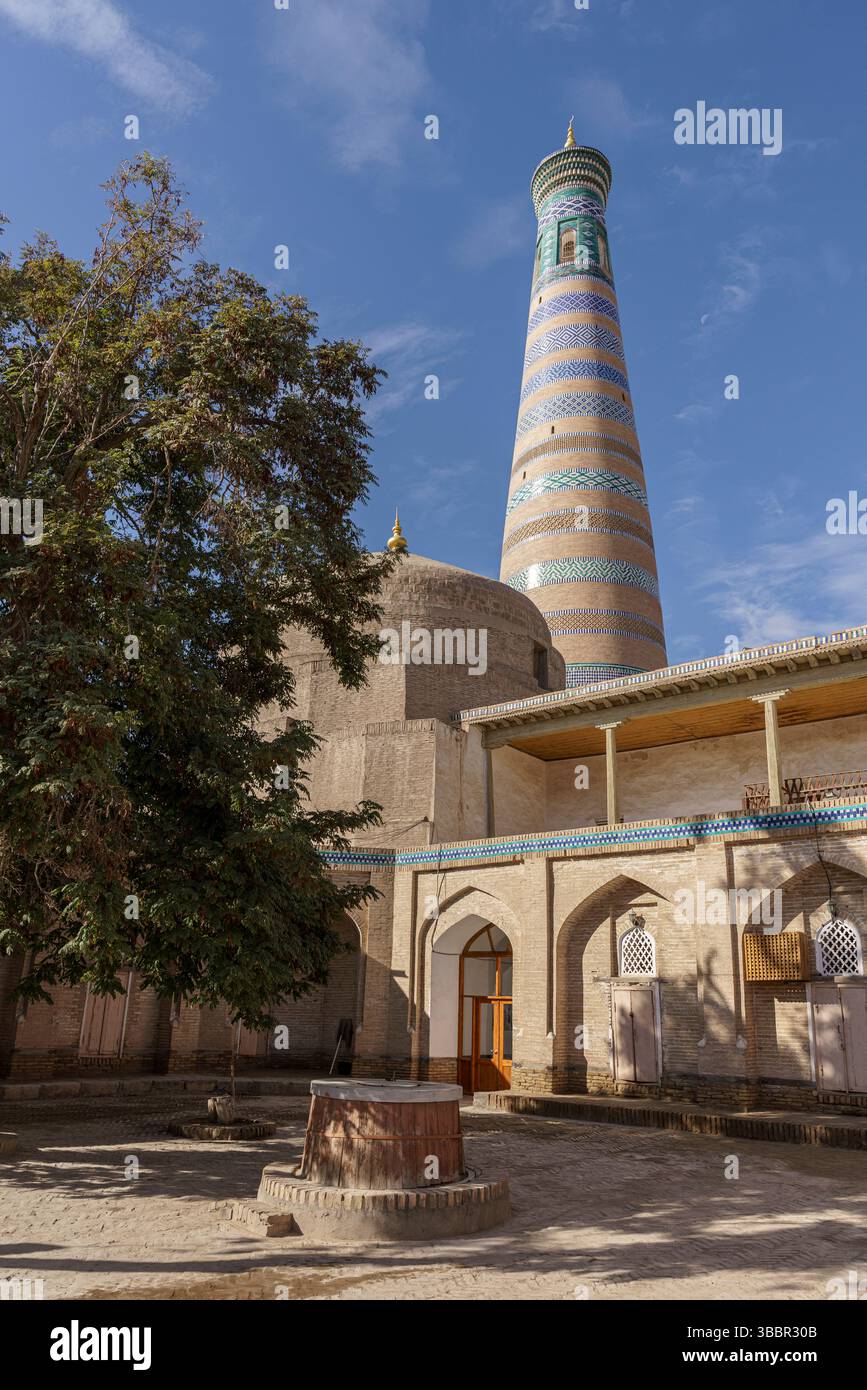 Un grand minaret avec des bandes turquoises se dresse à côté d'arbres feuillus et de murs sablonneux à Islam Khoja Madrasah à Khiva, Ouzbékistan. Banque D'Images