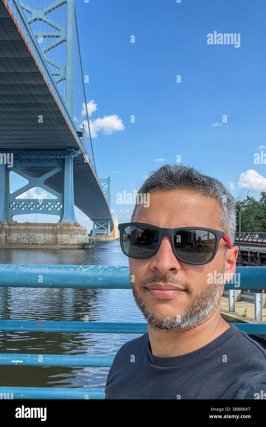 Un homme portant des lunettes de soleil est debout sur le pont Benjamin Franklin avec un ciel bleu en arrière-plan. Le pont est un pont suspendu, et l'eau b Banque D'Images