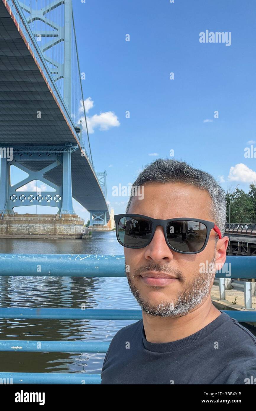 Un homme portant des lunettes de soleil est debout sur le pont Benjamin Franklin avec un ciel bleu en arrière-plan. Le pont est un pont suspendu, et l'eau b Banque D'Images