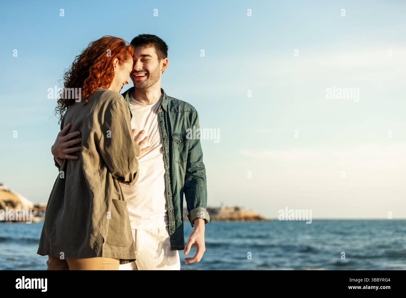 Heureux jeune couple partage un rire franc tout en embrassant sur la plage, baigné de lumière douce du soleil avec la mer et le ciel bleu en arrière-plan. Style de vie. Banque D'Images