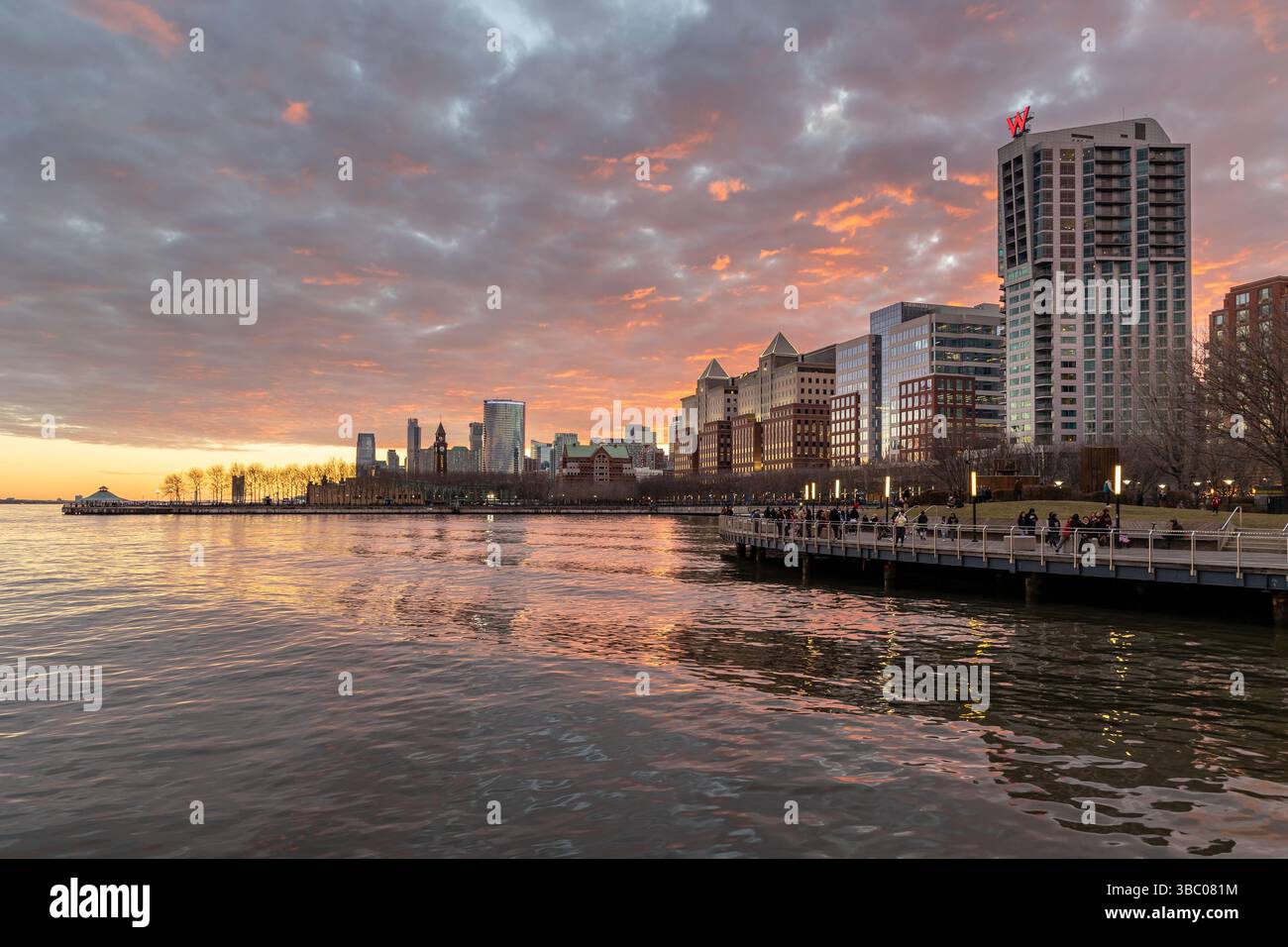Hoboken, NJ, États-Unis - 1er janvier 2023 : les gens se promènent le long du front de mer tandis que le coucher de soleil projette des couleurs éclatantes dans le ciel. Superbe horizon de la ville. Banque D'Images