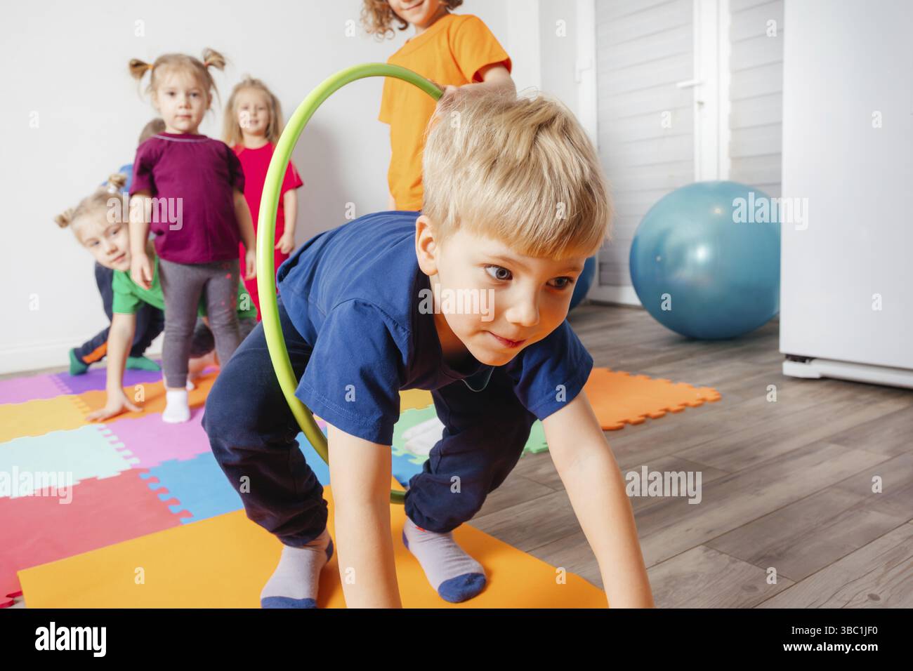 Enfant actif rampant sur des tapis colorés à travers des cerceaux de hula tenus par d'autres enfants. La compétition sportive à la maternelle Banque D'Images