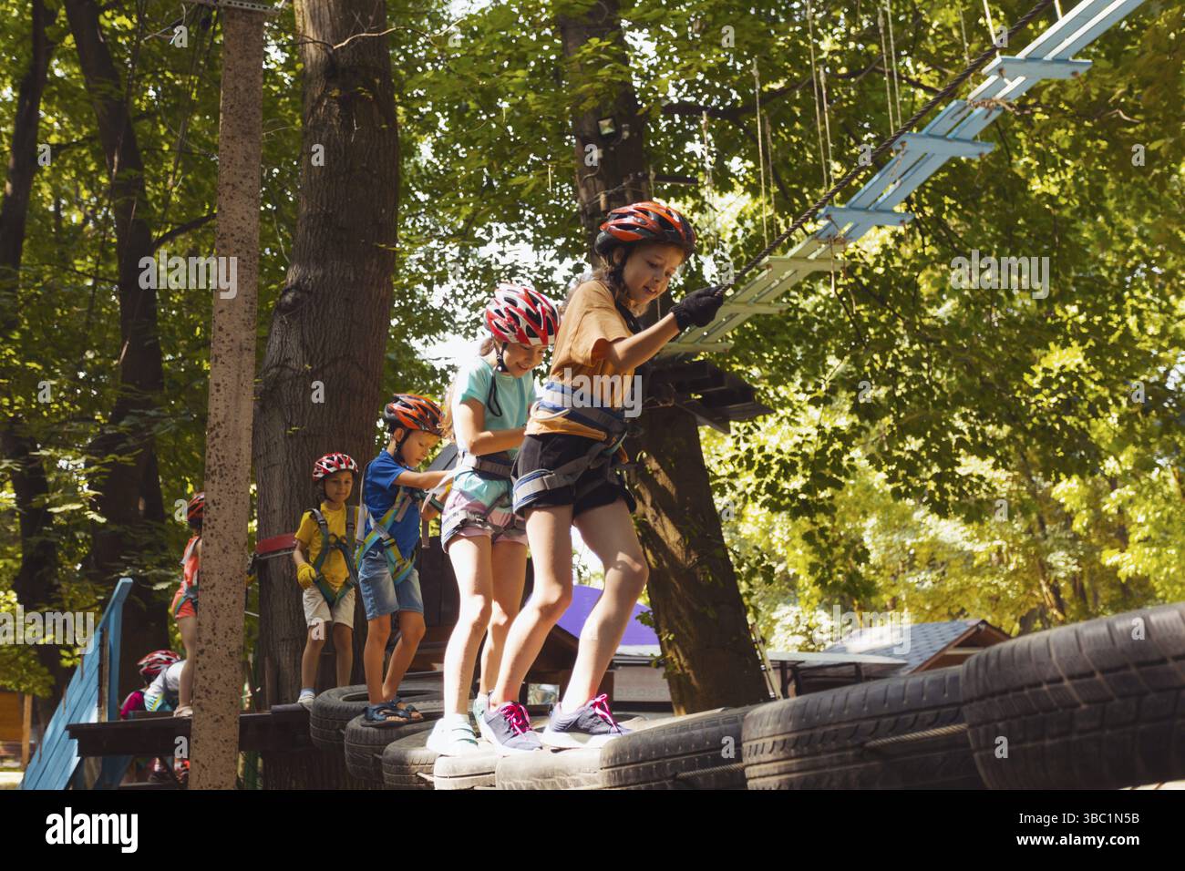 Vue arrière des enfants qui surmontent les obstacles dans le parc à cordes. Les enfants en casques passent devant l'arbre Banque D'Images