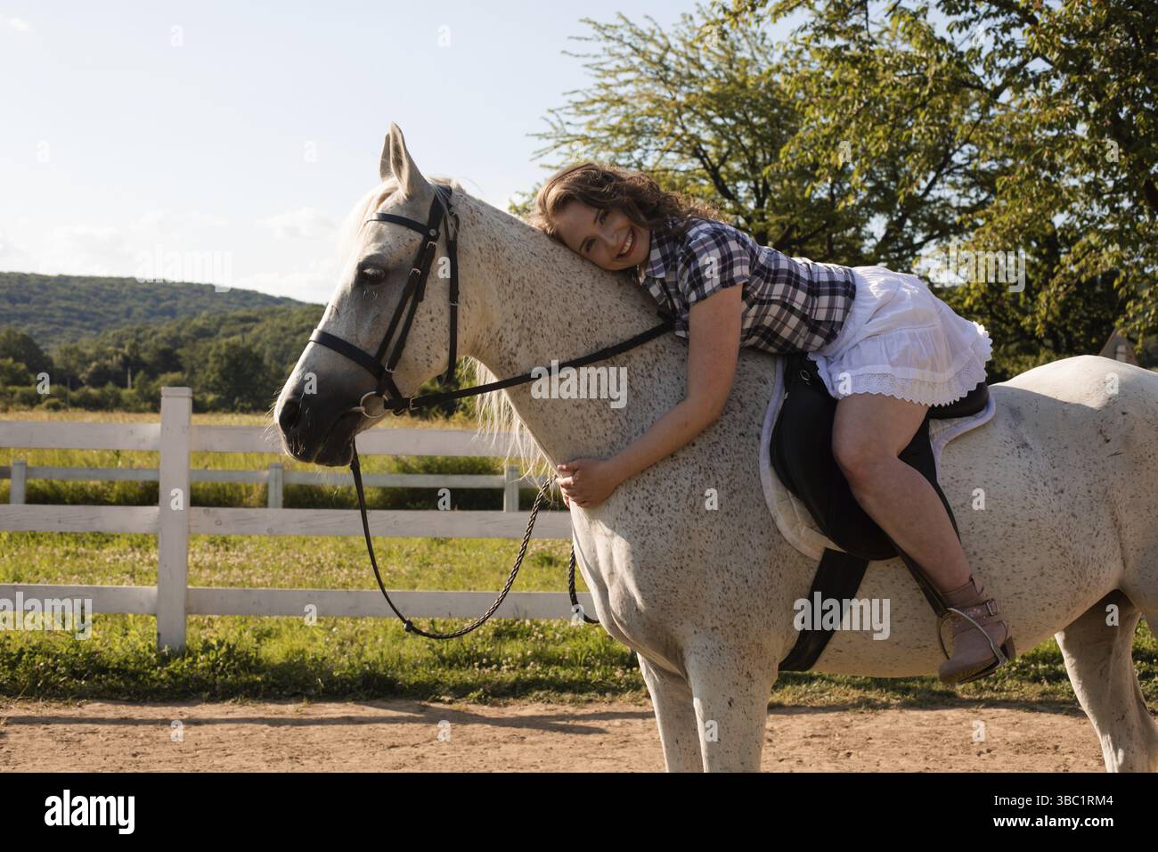 La femme adulte souriante en jupe blanche repose sur un cheval blanc.Femme avec un cheval sur un fond de belle nature Banque D'Images