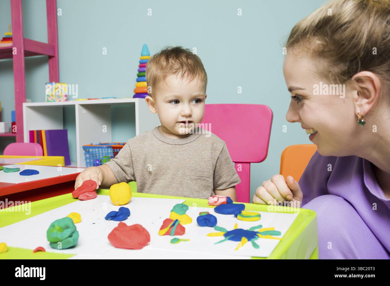 Le tout-petit garçon et la femme soignante se regardent. Le garçon sculpte utilise de la plasticine avec l'aide d'un tuteur Banque D'Images
