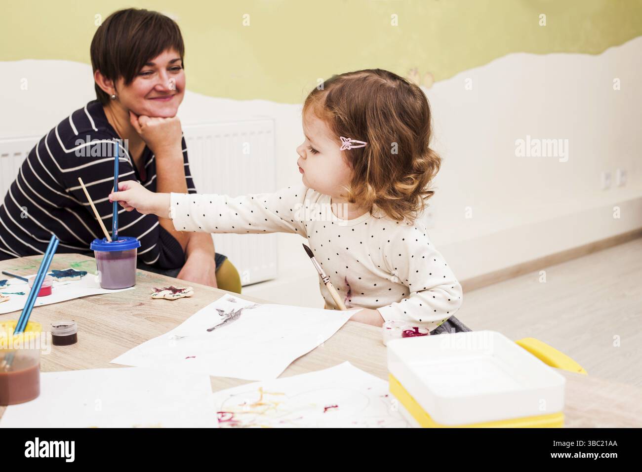 La fille peint avec un pinceau et des aquarelles au jardin d'enfants. Le professeur sourit Banque D'Images