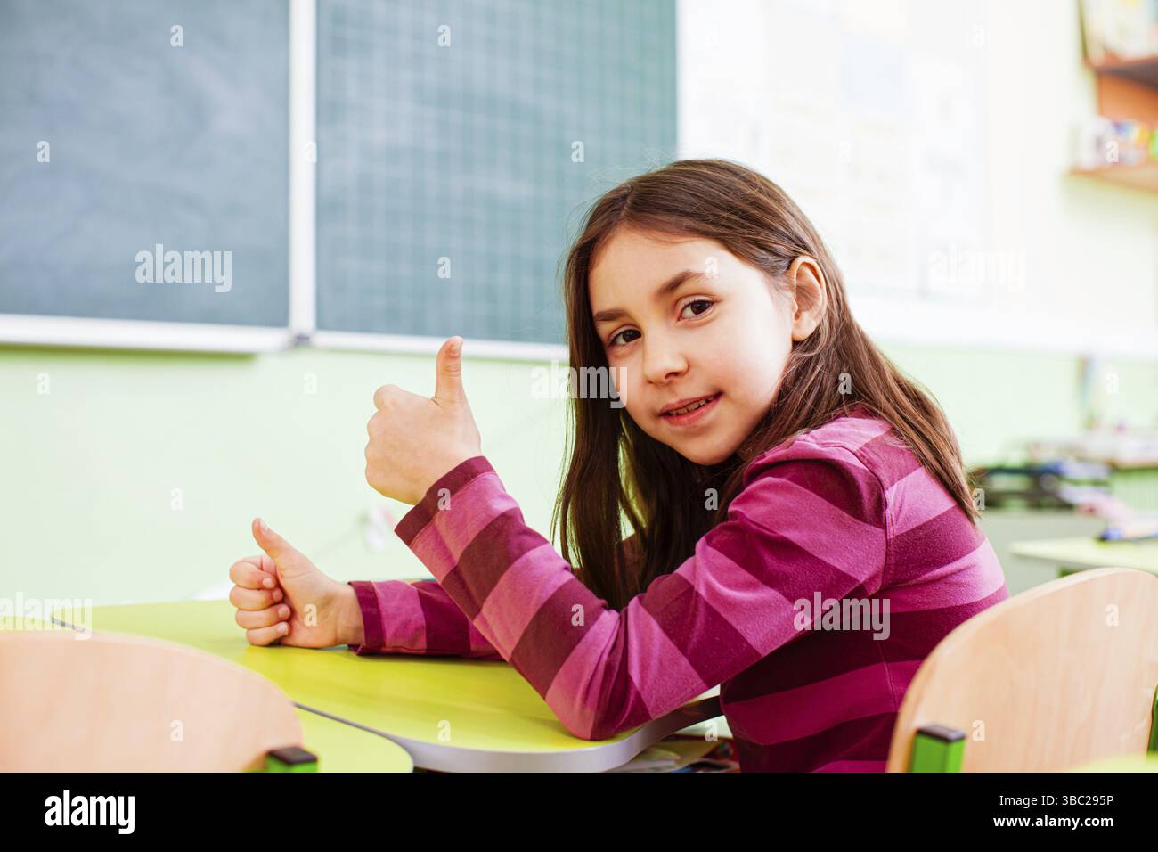 Portrait d'une fille de l'école primaire assise à son bureau avec le pouce vers le haut, souriant, regardant l'appareil photo. Fille avec le désir fort d'étudier appréciant son temps Banque D'Images