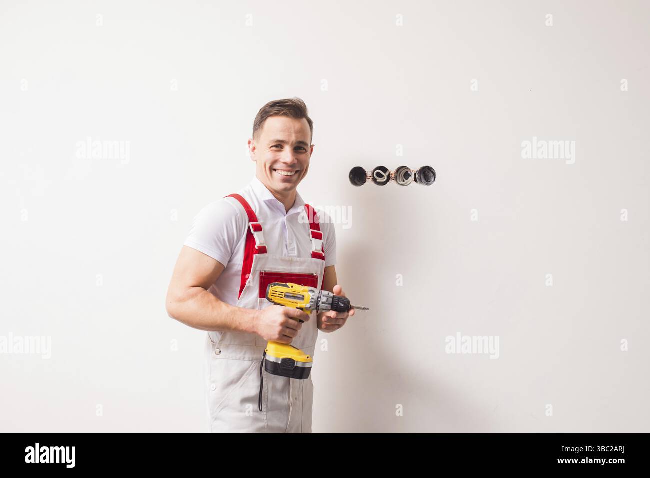 Le jeune électricien fort tient une perceuse. Il se tient aux prises contre le mur blanc en souriant et en regardant la caméra Banque D'Images