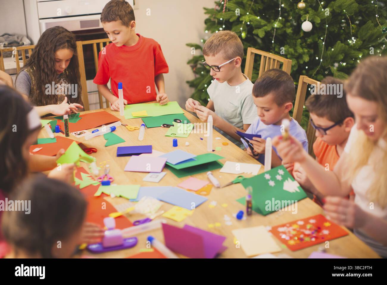 Grand groupe d'enfants de l'école primaire faisant des cartes de Noël pour leurs familles. Grande table en bois avec divers papiers colorés, colle, ciseaux. Sapin Banque D'Images