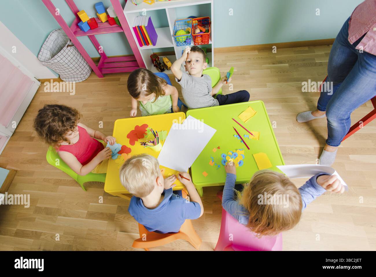 Cours d'art au jardin d'enfants, vue de dessus. Beaux enfants collant et coupant du papier sur des tables en plastique coloré. Concept de développement de l'enfant Banque D'Images