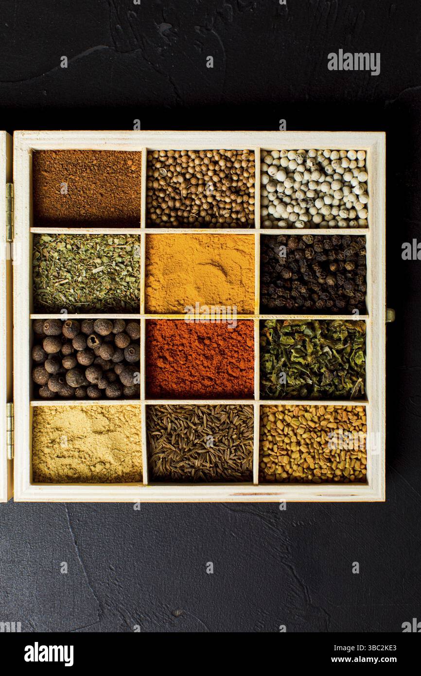 Rangement en bois pour les épices. Boîte divisée en cellules, remplie de diverses herbes sèches et épices. Condiments colorés et variés, vue du dessus sur le fond noir Banque D'Images