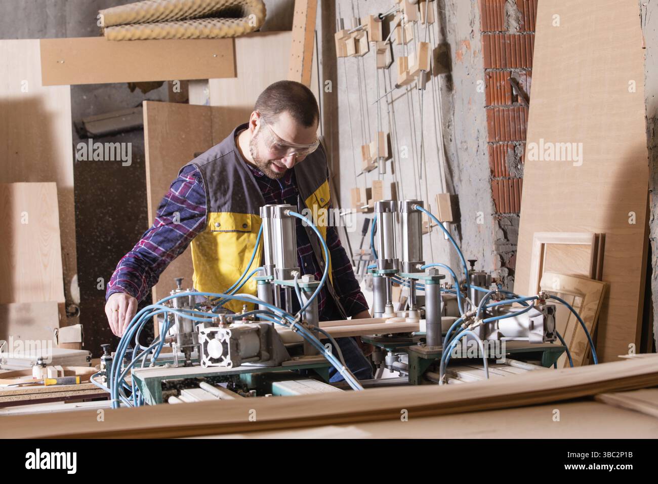 Portrait d'un jeune employé gai travaillant dans le bois utilisant une machine pneumatique dans un atelier poussiéreux. Production de meubles. Homme dans des lunettes de protection Banque D'Images