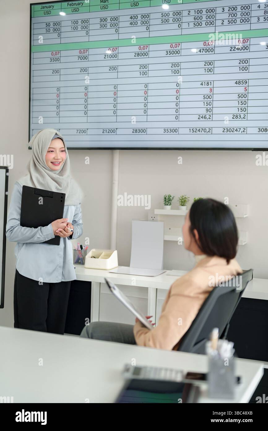 Deux femmes d'affaires dans un bureau analysant des données financières sur un écran. Une femme asiatique en hijab présente des données à un collègue. Discuter des tendances, des ventes, p Banque D'Images